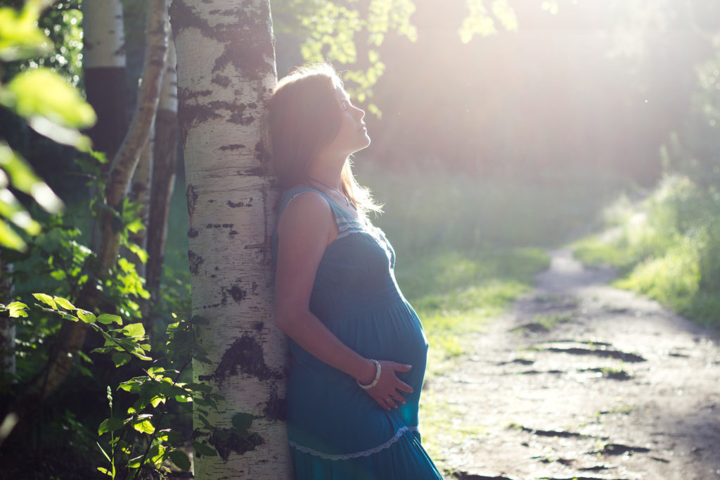Eine schwangere Frau lehnt sich gegen einen Baum und geniesst die Sonne.